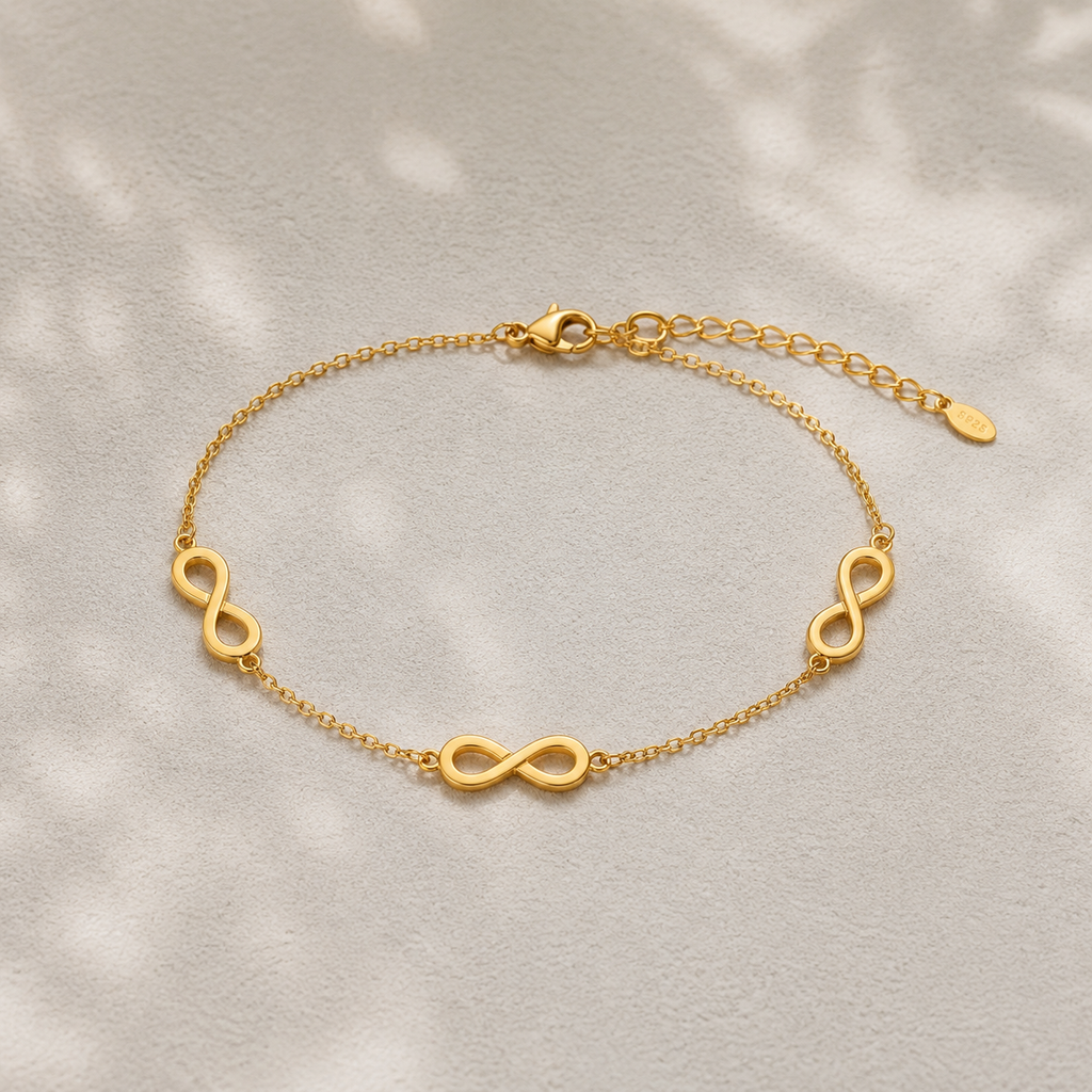 A delicate gold chain bracelet featuring three polished infinity symbols spaced evenly along the chain, displayed on a textured light gray surface with soft leaf shadows.