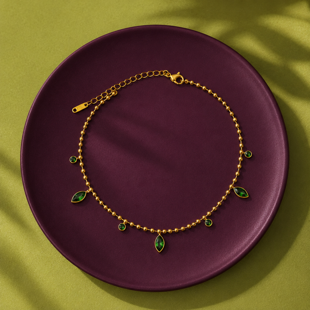 A gold chain anklet with green marquise and round gemstones, displayed on a dark purple circular plate over an olive green background with soft leaf shadows.