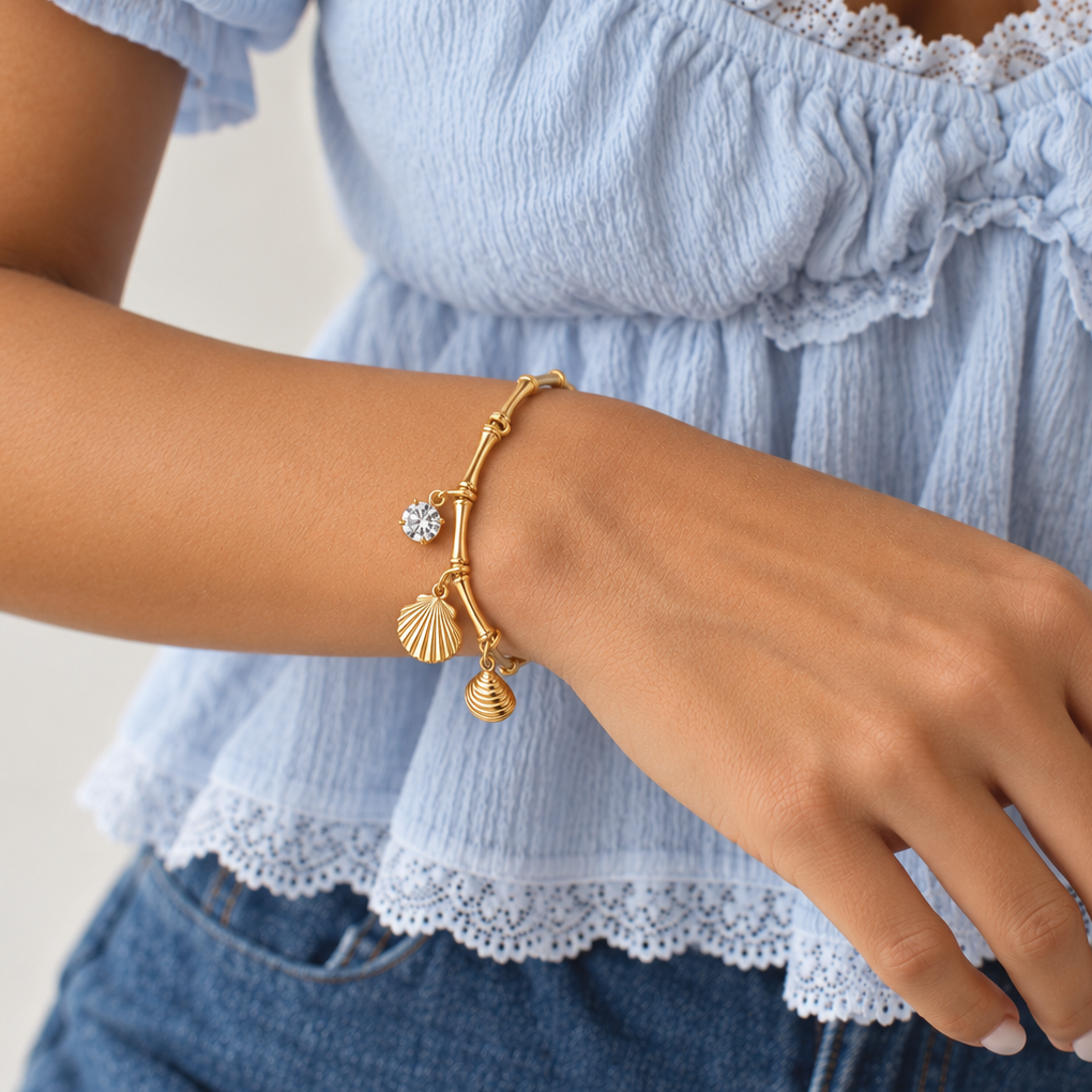 A gold charm bracelet with a bamboo-style link chain, a small flower charm, and two seashell charms, shown worn on a woman's wrist.