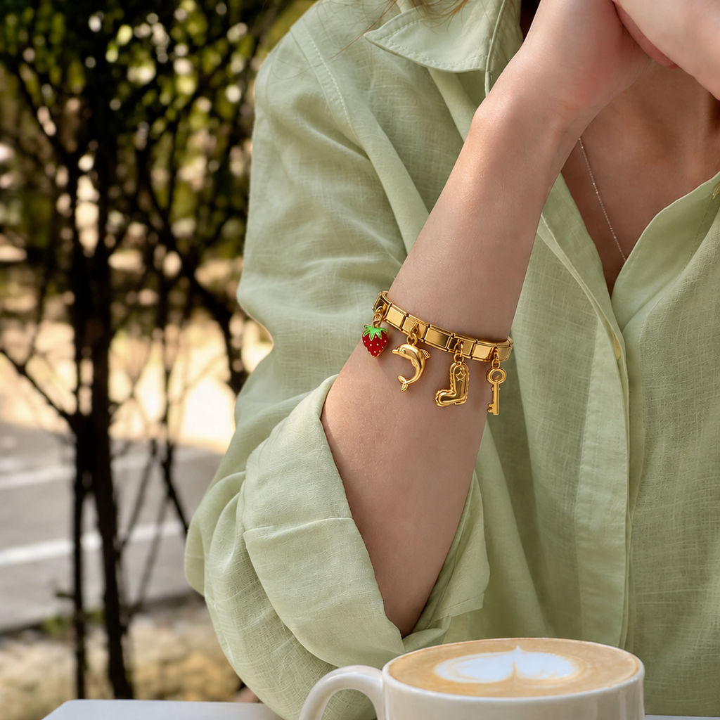 A close-up of a person wearing a gold charm bracelet on their wrist, styled with a light green linen shirt. The bracelet features charms including a strawberry, a horseshoe, a boot, and a key.