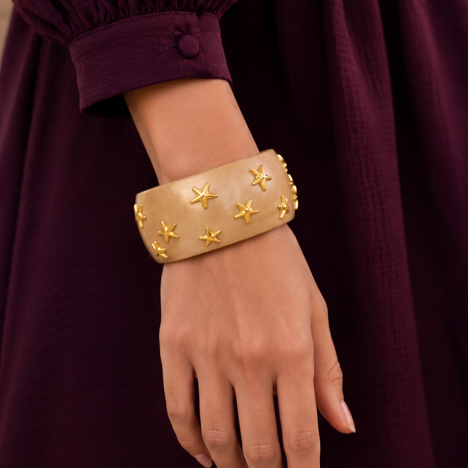A close-up of a person's hand and wrist wearing a wide beige cuff bracelet decorated with small gold star charms, set against a dark burgundy dress.
