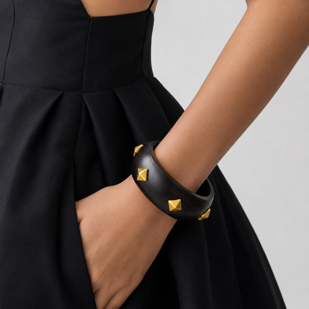 A close-up of a model's arm wearing a wide black leather cuff bracelet adorned with gold pyramid studs, paired with a black pleated dress against a light gray background.