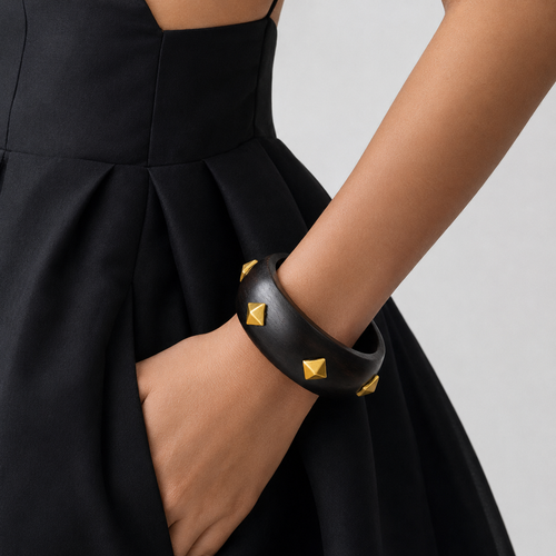 A close-up of a model's arm wearing a wide black leather cuff bracelet adorned with gold pyramid studs, paired with a black pleated dress against a light gray background.