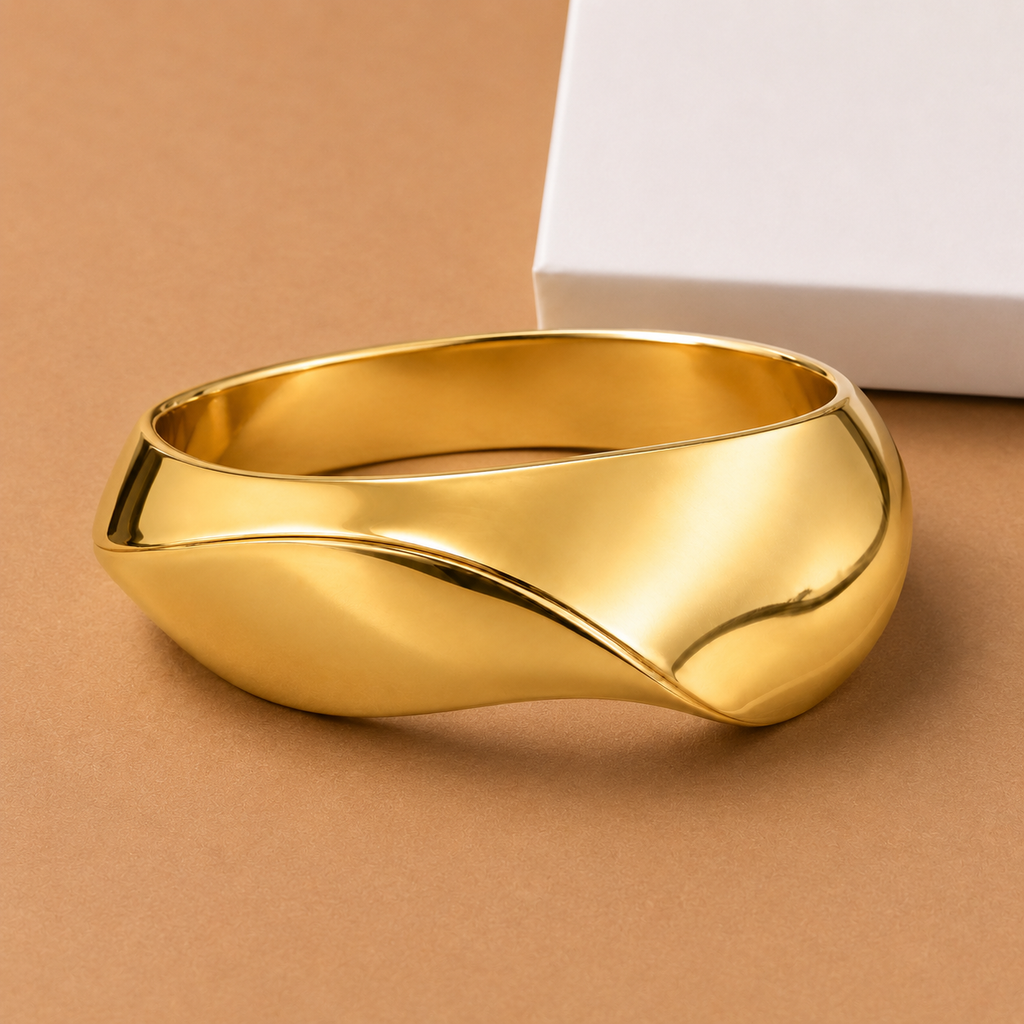 A polished gold bangle bracelet with a wavy, fluid organic shape, displayed on a warm brown background next to a white box.