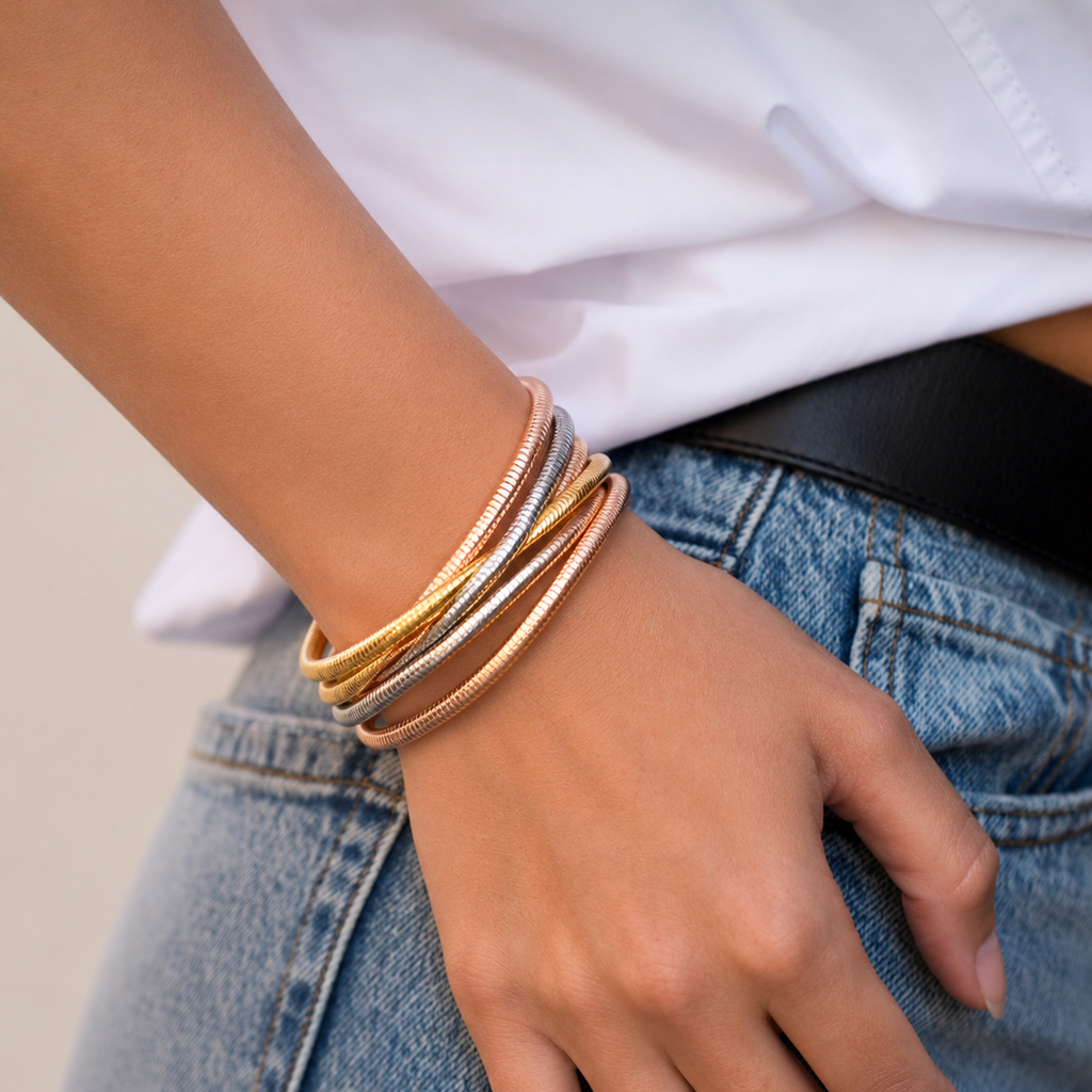 A close-up of a person's wrist wearing a stack of four textured bangle bracelets in gold, rose gold, and silver tones, styled with a white shirt and blue jeans.