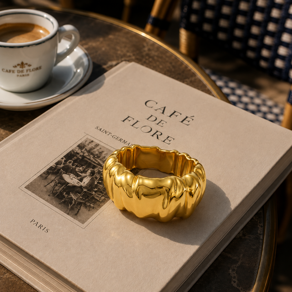 A chunky, highly polished gold ring with a wavy, pleated texture rests on the edge of an open book. The book, titled 'Café de Flore', is placed on a round wooden table alongside a matching coffee cup and a patterned chair in the background, creating a warm, cafe-themed lifestyle setting.
