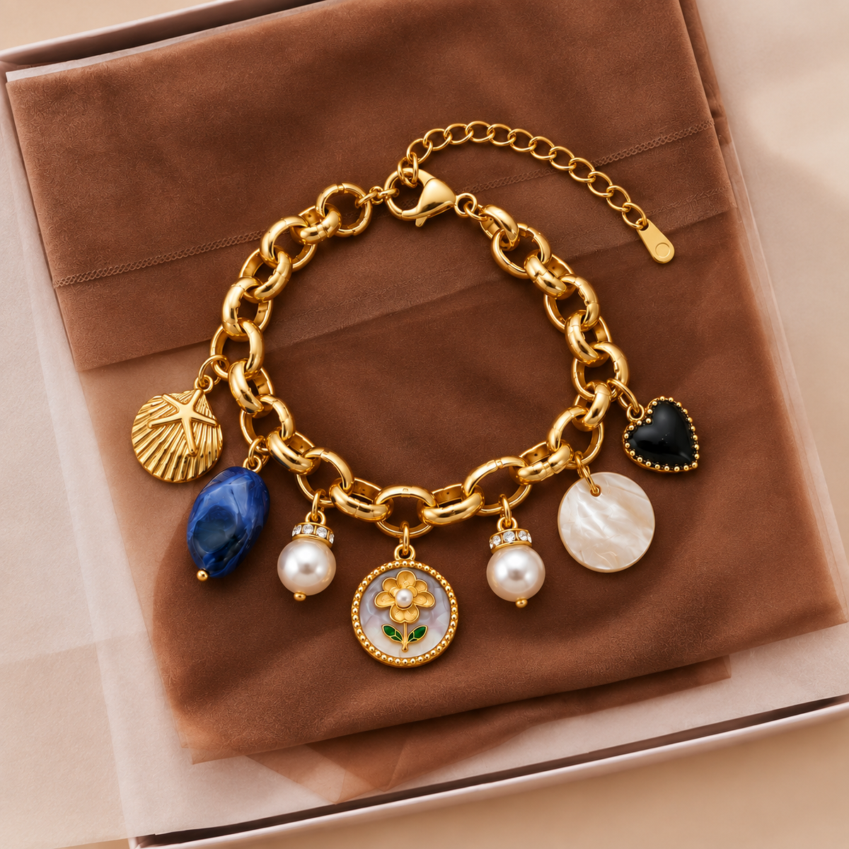 A gold-tone charm bracelet displayed on a brown velvet jewelry box. The bracelet features a chunky link chain with multiple charms, including a blue faceted stone, a white pearl, a white mother-of-pearl disc, a gold flower with green leaves, a black heart, and a gold starfish shell.