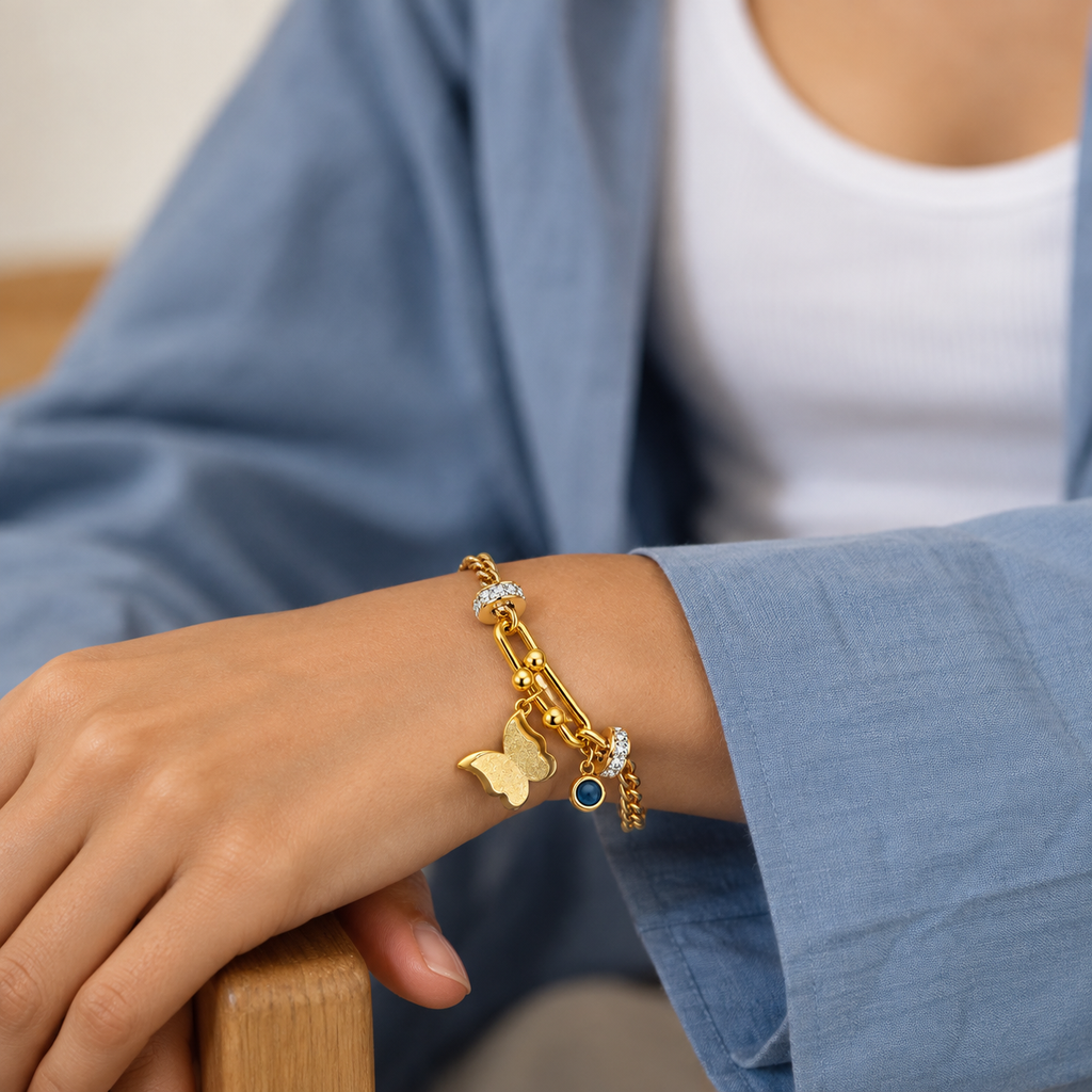 A gold charm bracelet with a butterfly, safety pin, and small blue gemstone charm, shown worn on a person's wrist.