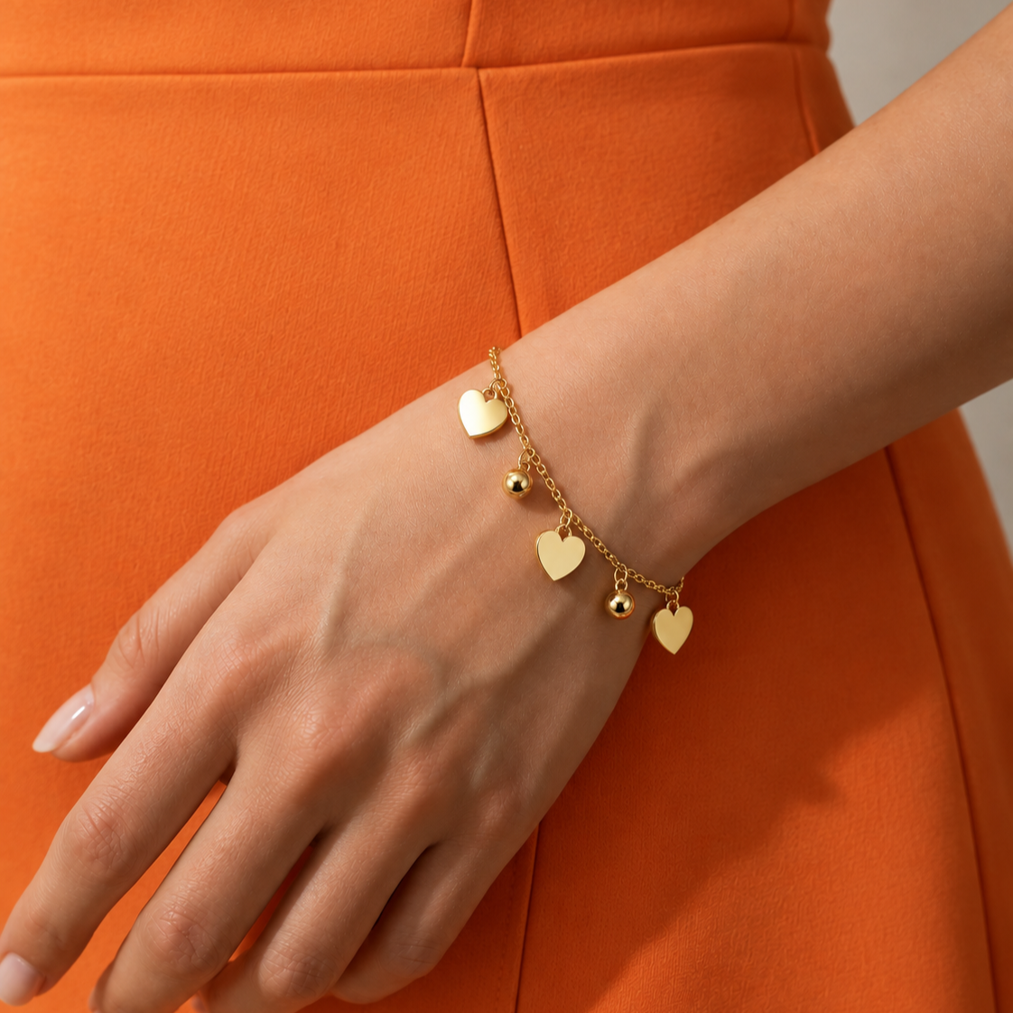 A delicate gold chain bracelet with three small heart charms and a tiny bead charm, worn on a woman's wrist. The person is wearing a bright orange dress, and the background is a soft, out-of-focus neutral gray.