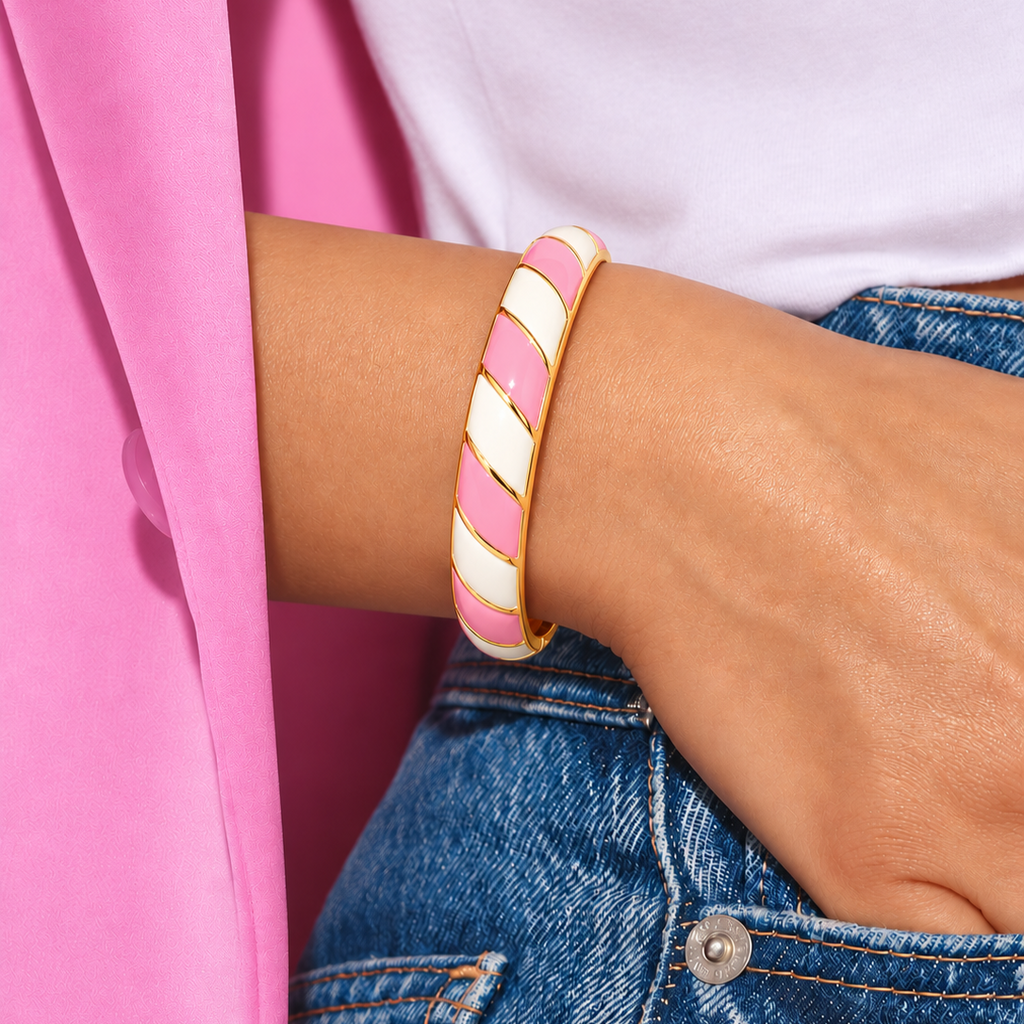 A close-up of a person's wrist wearing a gold-plated bangle bracelet with alternating pink and white diagonal stripes, styled with a white top and blue denim jeans.