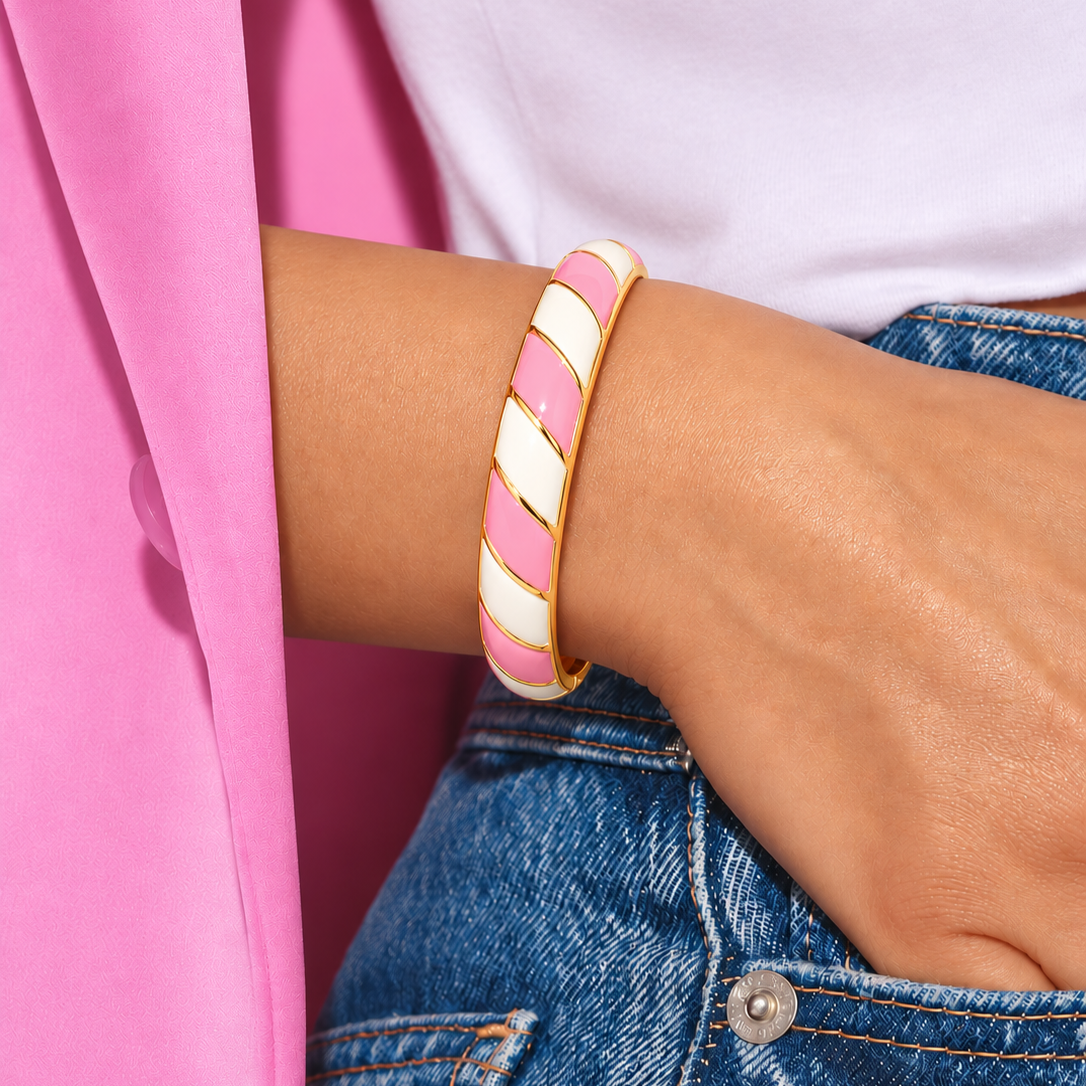 A close-up of a person's wrist wearing a gold-plated bangle bracelet with alternating pink and white diagonal stripes, styled with a white top and blue denim jeans.