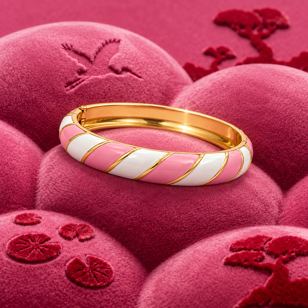 A gold-plated bangle bracelet with alternating pink and white striped enamel segments, resting on a textured pink velvet surface with embroidered motifs.