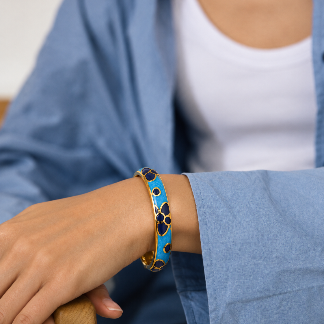 A close-up of a person wearing a gold bangle bracelet with a bright blue enamel body and dark floral accents, styled with a white tank top and a light blue shirt.