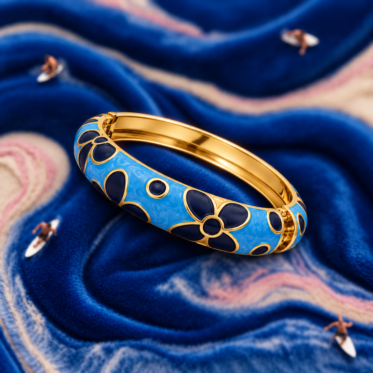 A gold-plated bangle bracelet with a bright blue enamel background and dark navy floral motifs outlined in gold, displayed on a textured blue velvet surface.