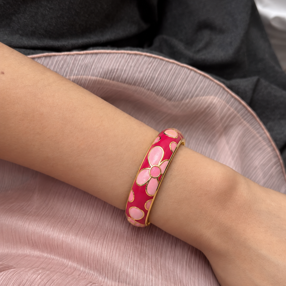 A close-up of a person's wrist wearing a pink and gold floral enamel bangle bracelet, resting on a pink satin fabric.