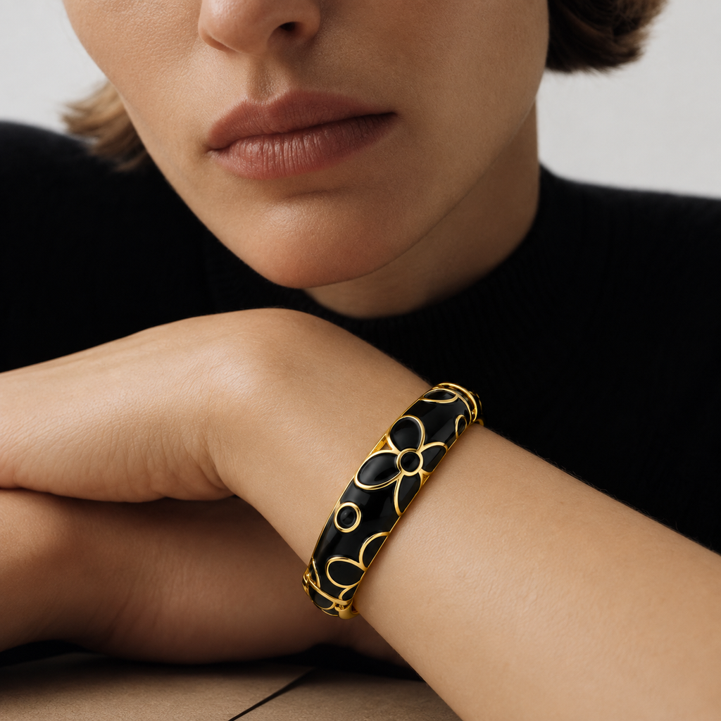 A close-up of a model's wrist wearing a black and gold bangle bracelet with a floral pattern, set against a dark sweater and neutral background.