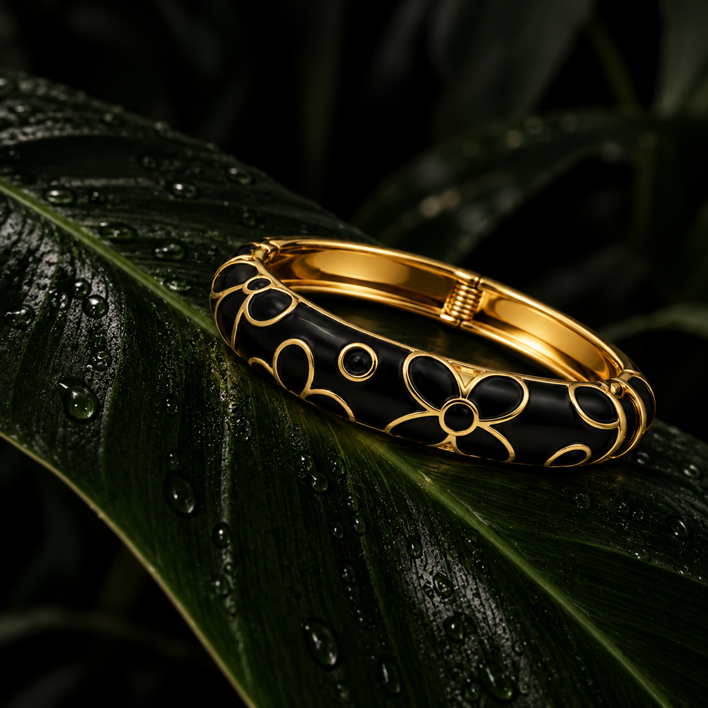 A gold and black bangle bracelet with a floral pattern, resting on a glossy, water-droplet-covered green leaf against a dark background.