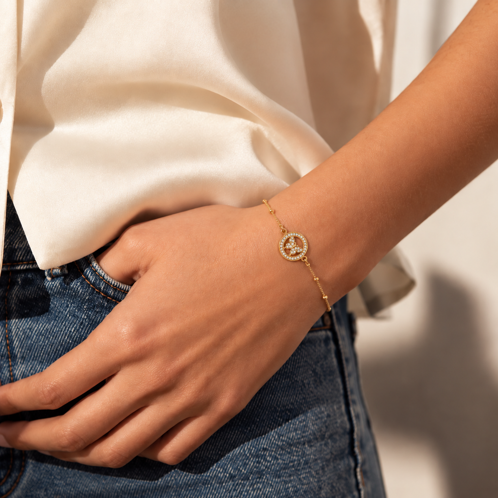A close-up of a person's wrist wearing a delicate gold chain bracelet with a circular crystal-encrusted pendant featuring a central letter 'A'. The person is wearing a cream satin blouse and blue jeans, with warm sunlight casting soft shadows.