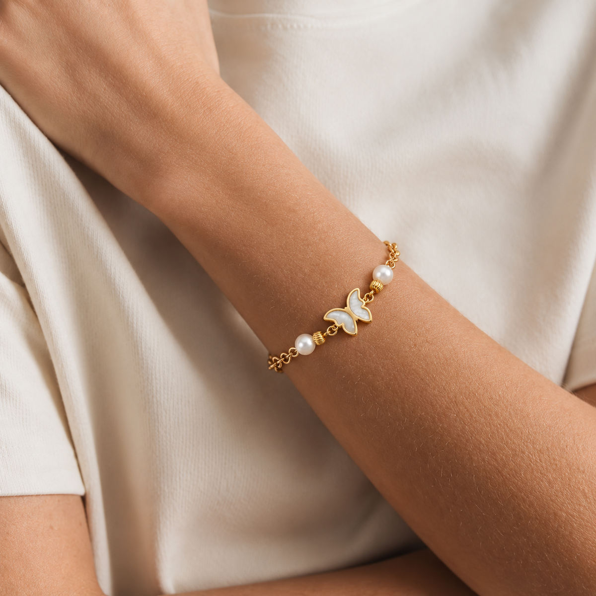 A delicate gold chain bracelet with a white mother-of-pearl butterfly charm and pearl beads, shown worn on a person's wrist against a soft cream-colored top.