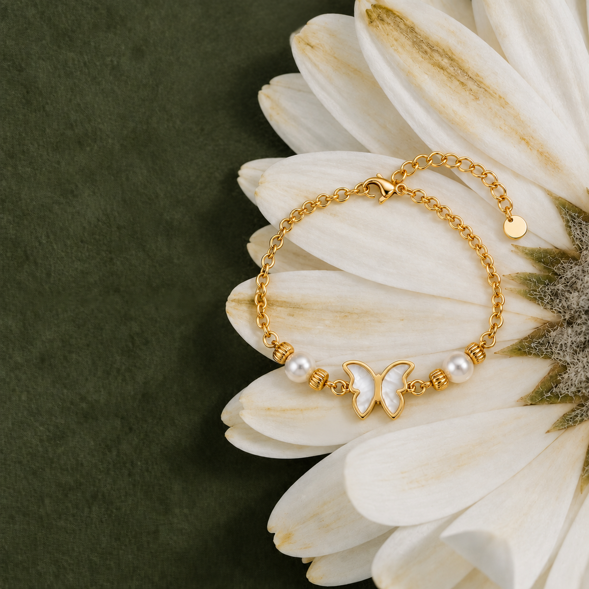 A gold chain bracelet with a butterfly charm, white pearls, and gold spacer beads, styled on the petals of a white daisy flower against a dark olive green background.