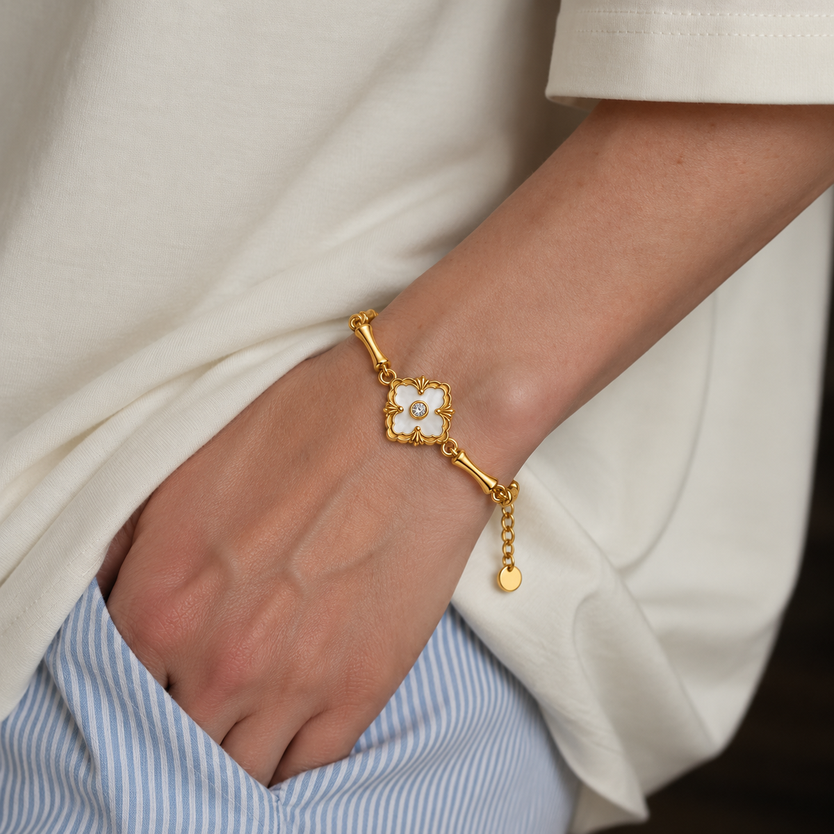 A close-up of a person's wrist wearing a delicate gold bracelet with a white flower charm and a small pearl center, styled with a cream-colored top and blue striped pants.