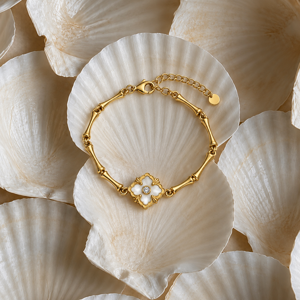 A gold bracelet with a central flower motif featuring white pearl-like accents, displayed on a large white clam shell among other shells.