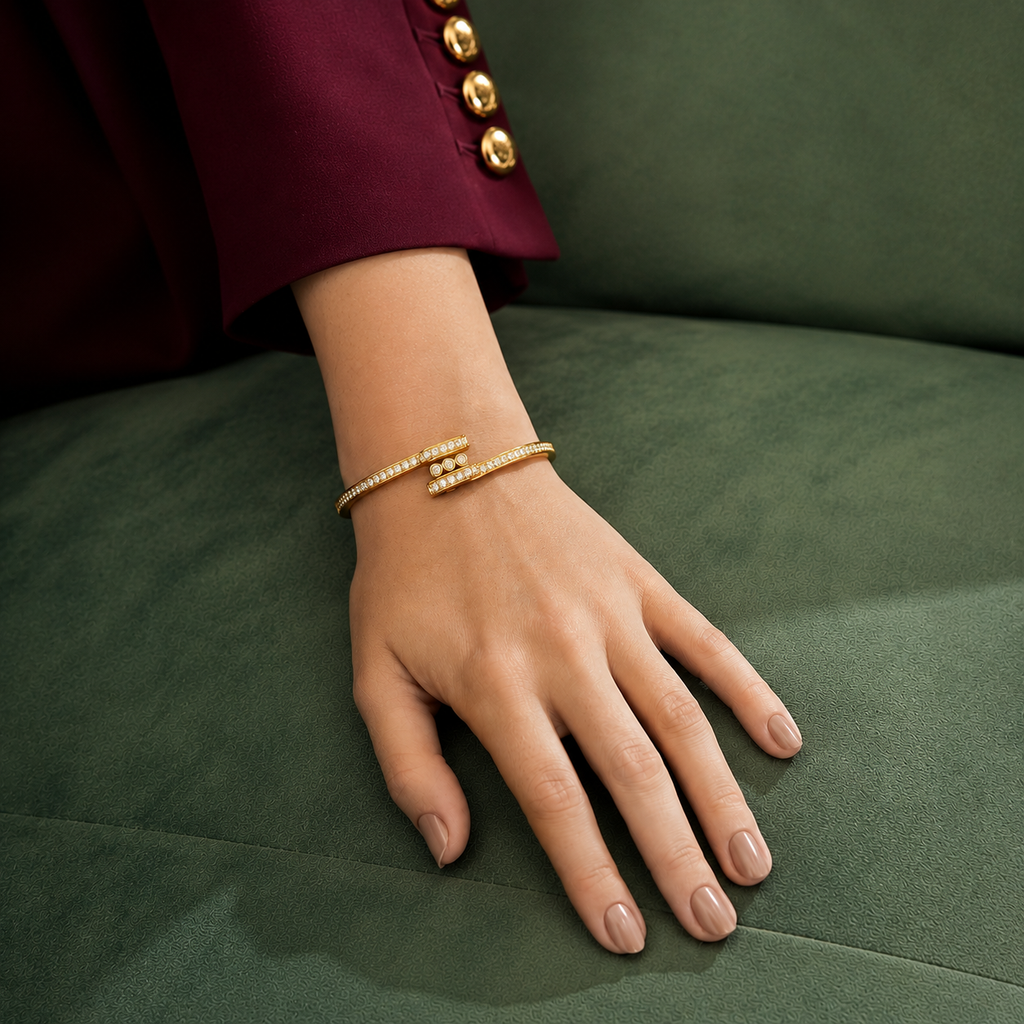 A close-up of a person's hand resting on a dark green velvet surface, wearing a delicate gold wrap bracelet adorned with small clear stones. The person is wearing a deep burgundy sleeve with gold buttons.