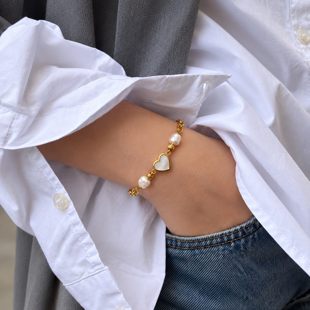 A gold bracelet with white pearl-like beads and a heart-shaped charm, worn on a person's wrist. The person is wearing a white button-up shirt and blue jeans, with a gray blazer draped over their shoulder.