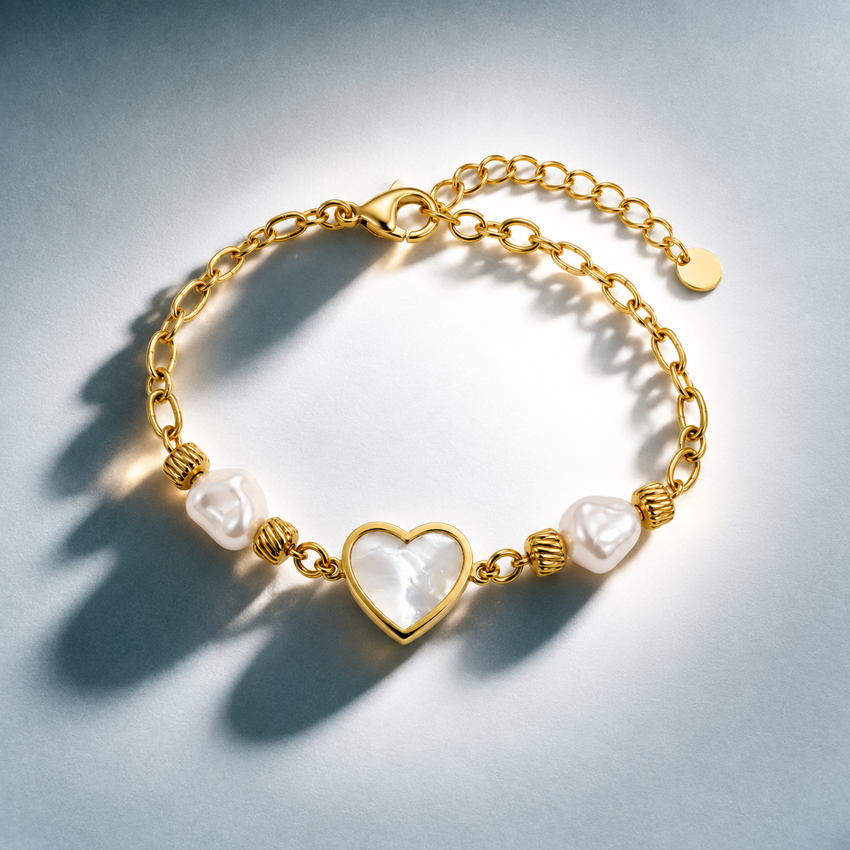 A gold chain bracelet featuring a central heart-shaped pearl with a gold border, flanked by two textured gold beads and a larger irregular white pearl, photographed on a light gray background with soft shadows.