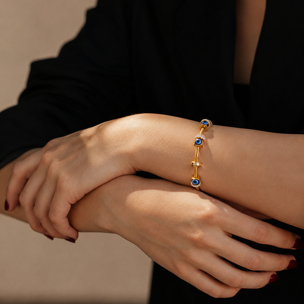 A close-up of a person's hands and arms wearing a delicate gold bracelet with multiple oval blue gemstone links, styled with a black top against a neutral beige background.