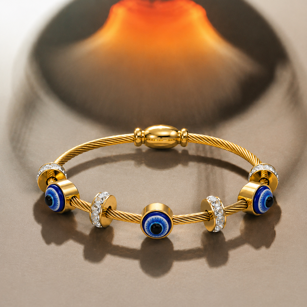 A gold cable bracelet with evil eye charms and clear crystal accents, displayed on a reflective beige surface with a soft blurred orange background.