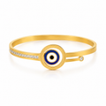 Gold bangle bracelet with a central evil eye motif in blue, white, and black, accented by a row of small clear stones on one side and a single stone on the other, shown on a white background.