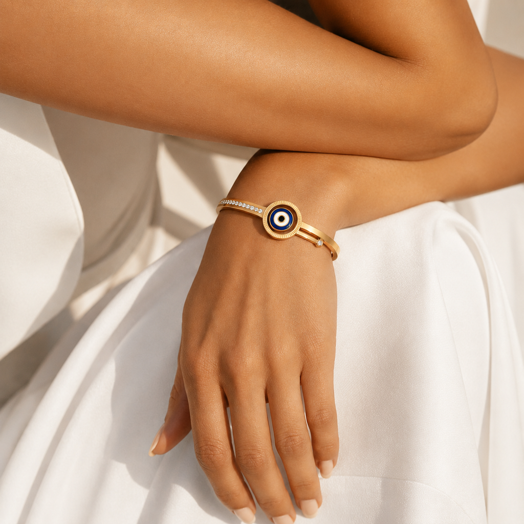 A gold-toned cuff bracelet with an evil eye motif and a row of small clear stones, worn on a model's wrist against a white draped fabric background.