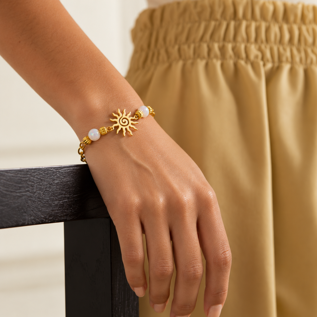 A gold bracelet with a sun charm and white beads worn on a person's wrist, shown against a beige skirt and dark wooden table.