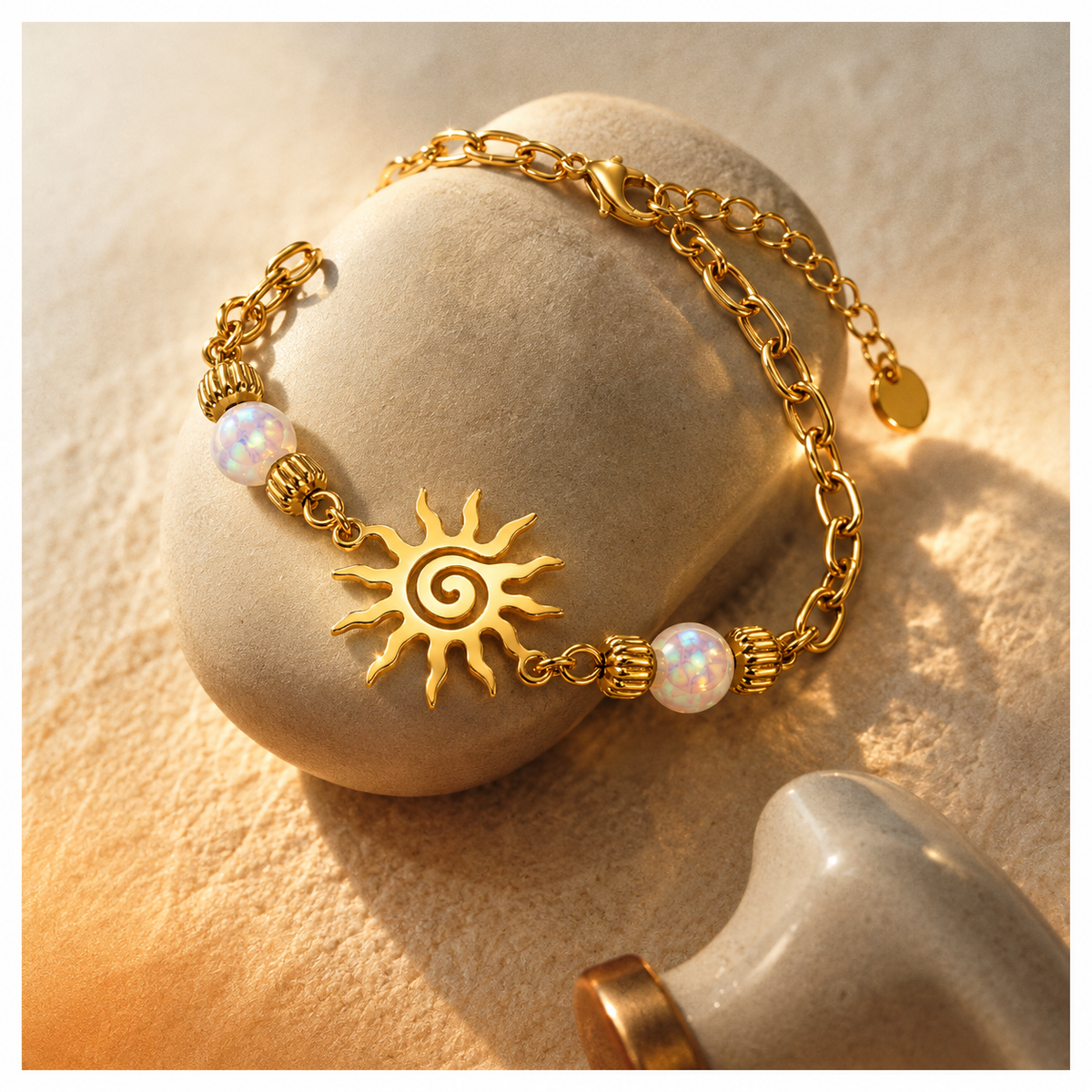 A gold-tone chain bracelet with a sun face charm, textured bead details, and white iridescent opal-like beads, styled on a smooth stone against a warm sandy background.