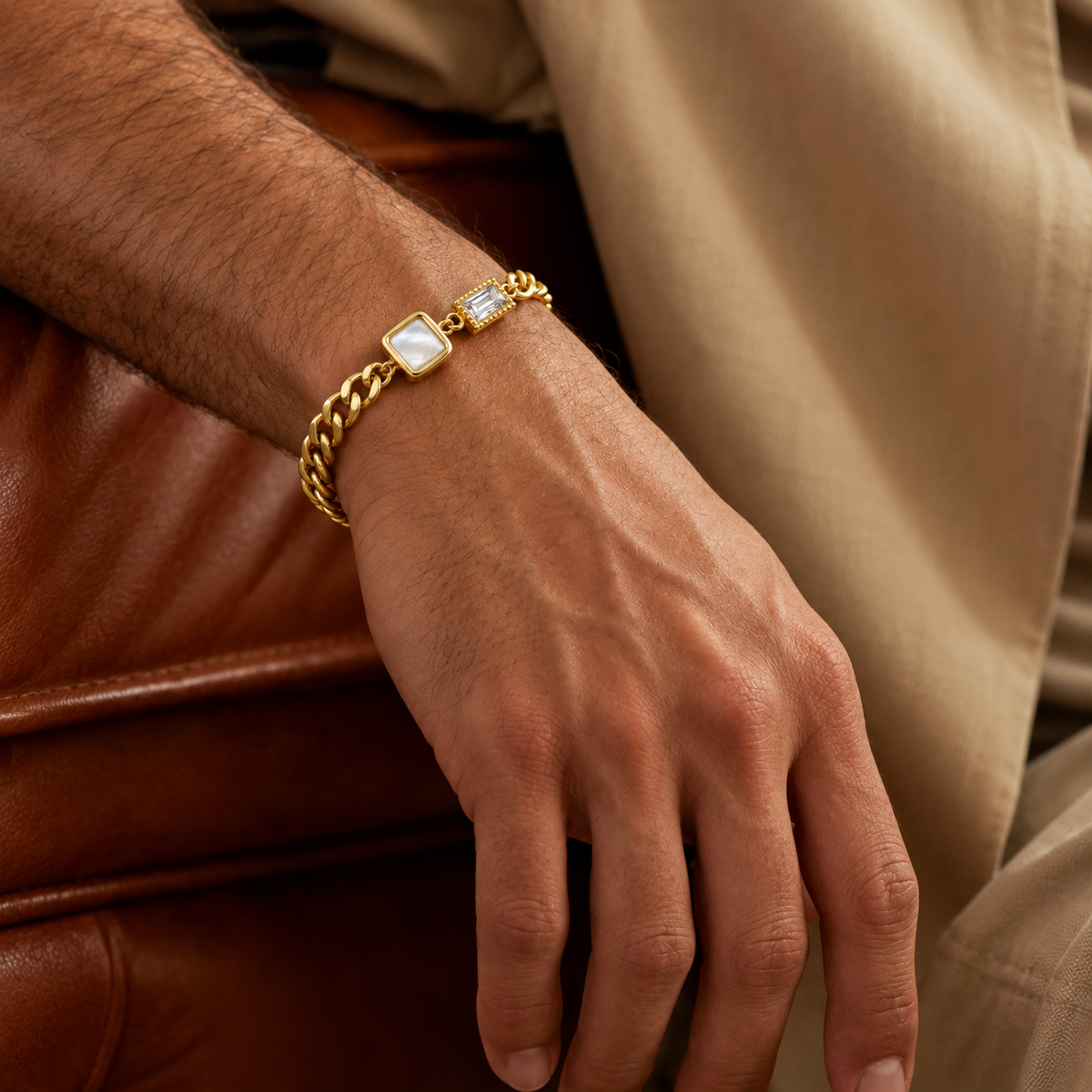 A close-up of a man's hand wearing a gold-toned chain bracelet with a rectangular mother-of-pearl inlay and a small clear stone, set against a background of brown leather and beige clothing.