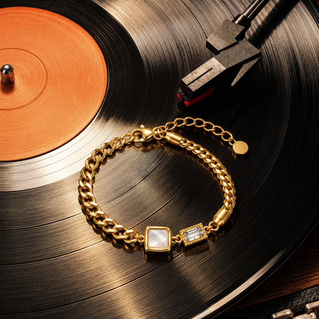 A gold chain bracelet with two square charm pendants, one featuring a white mother-of-pearl center and the other a clear rhinestone, displayed on a black vinyl record with a copper center label.
