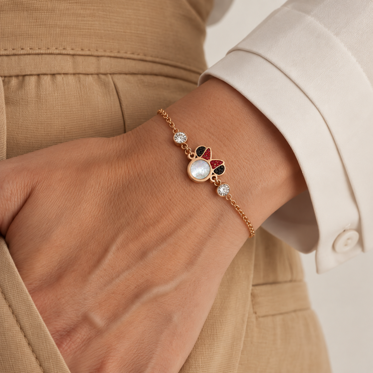 A close-up of a delicate gold chain bracelet worn on a wrist. The bracelet features a central oval white pearl-like stone, a small red and black enamel motif, and two clear crystal accents.