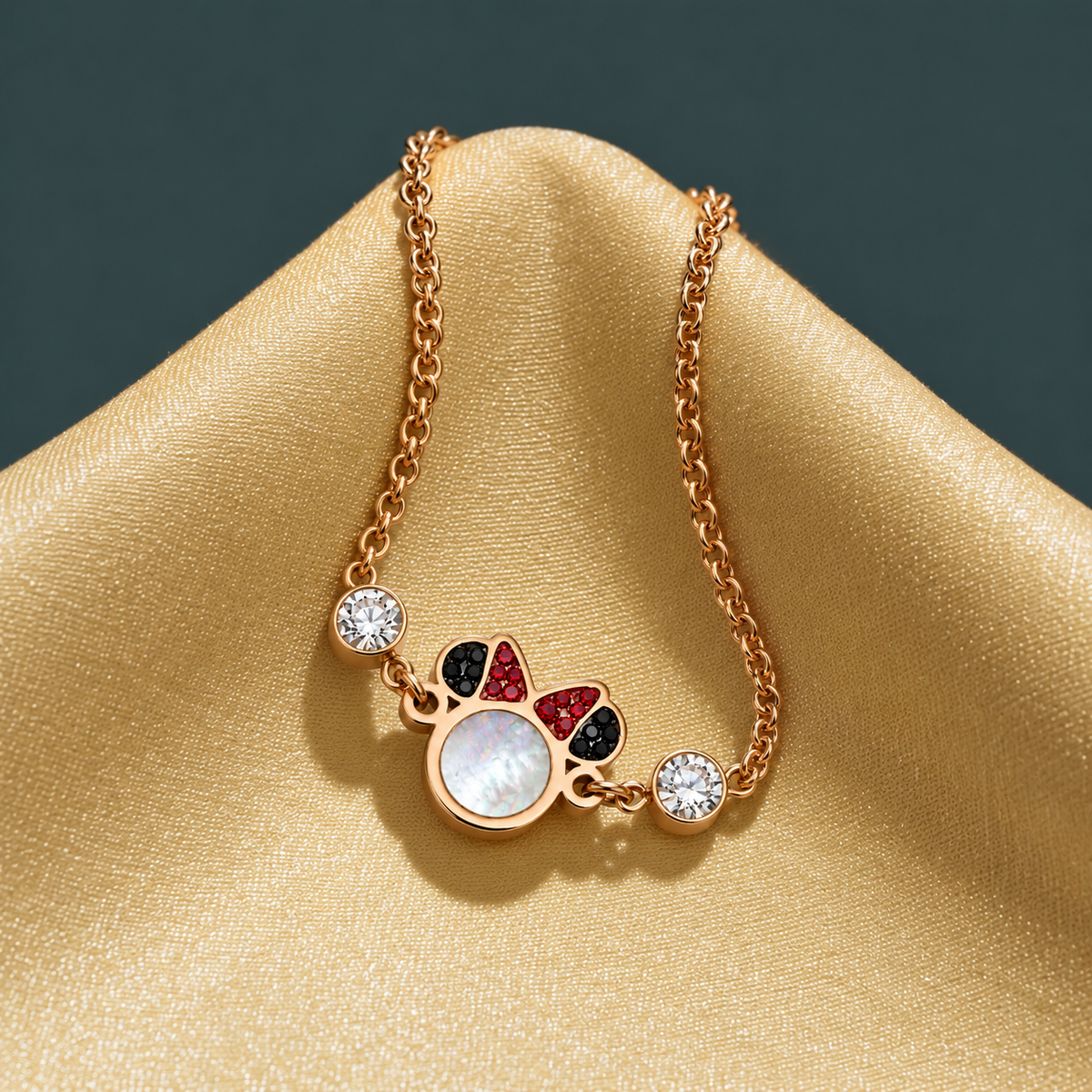 A rose gold chain necklace with a cat-shaped pendant featuring a white mother-of-pearl face, red and black eyes, and two clear rhinestone accents, displayed on a gold satin fabric background.