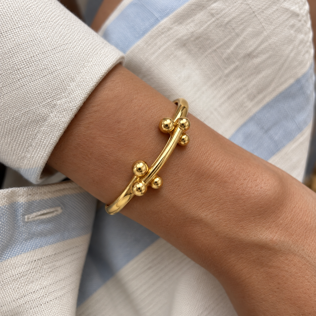 A close-up of a gold bangle bracelet with a curved bar and small bead-like spheres, worn on a person's wrist. The person is wearing a light-colored, textured shirt with blue stripes.
