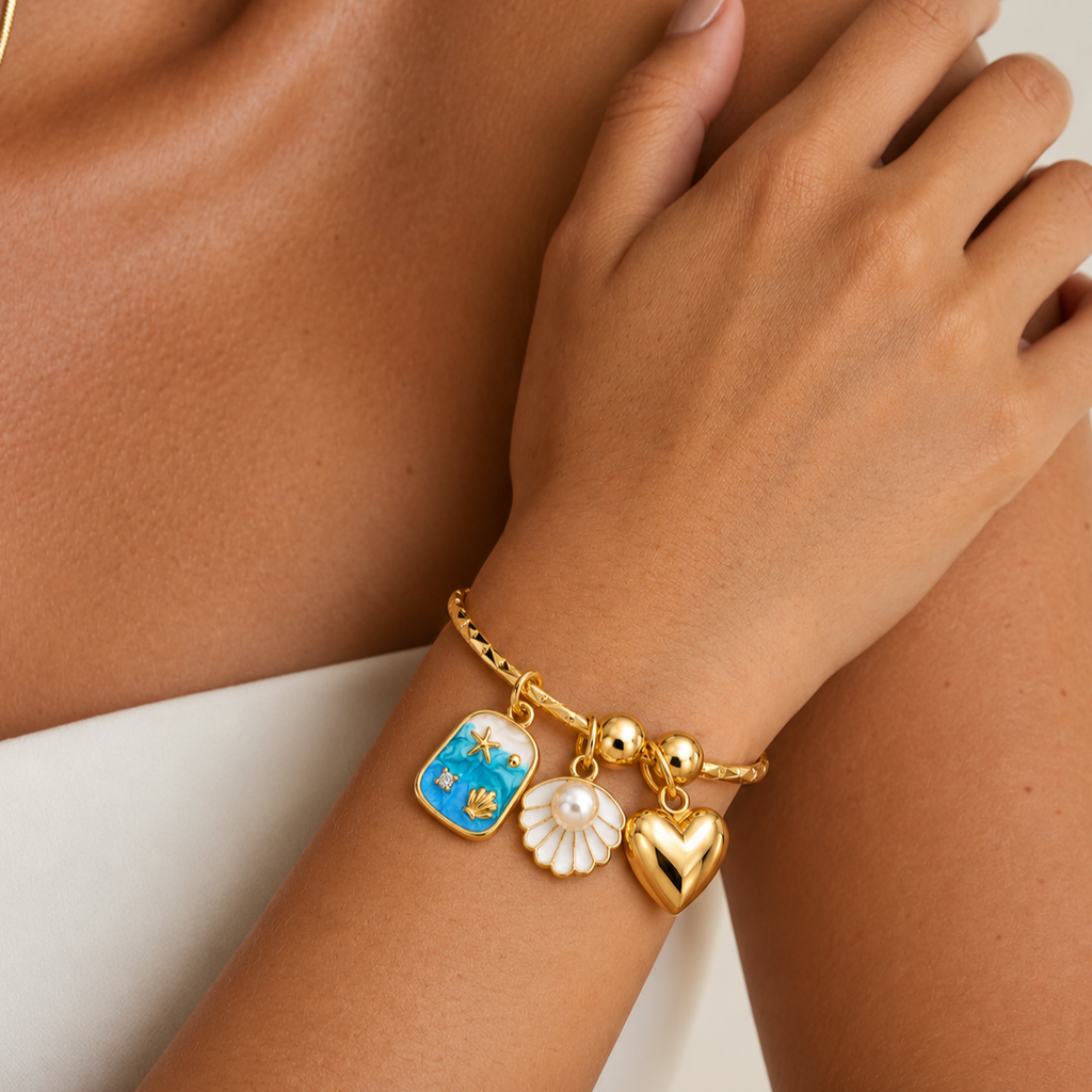 A close-up of a gold charm bracelet worn on a wrist. The bracelet features three charms: a blue and gold fish-shaped charm, a white flower charm with a pearl center, and a polished gold heart charm.