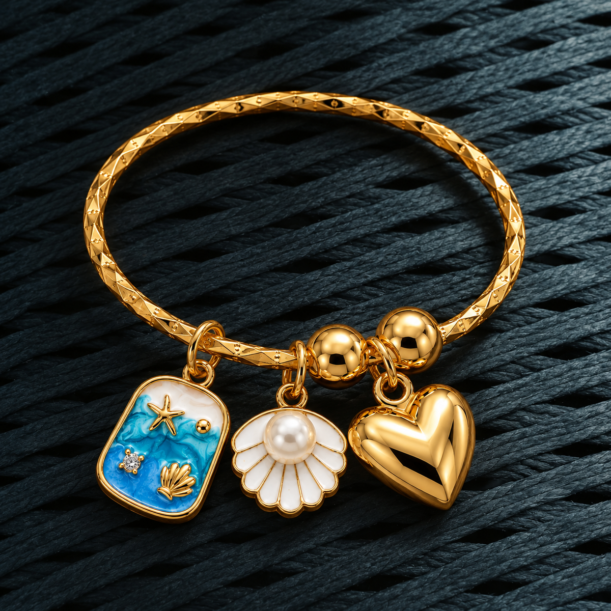 A gold-plated bangle bracelet with three charms: a blue enamel shell charm with gold starfish and seashell details, a white seashell charm with a pearl center, and a polished gold heart charm, displayed on a dark textured background.