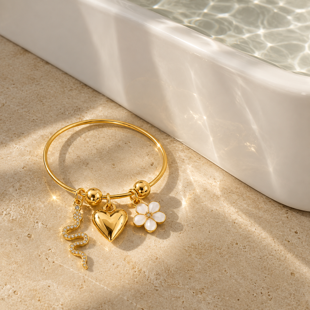 A gold-plated bangle bracelet with three charms (a heart, a flower, and a snake) resting on a beige stone surface next to a white ceramic tub with sunlight reflecting off it.