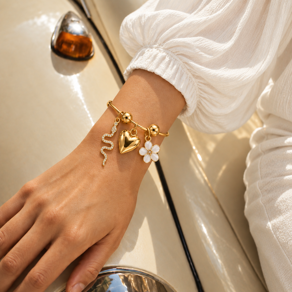 A close-up of a person's wrist wearing a delicate gold charm bracelet with a heart, flower, and snake charms, styled with a white long-sleeve blouse next to a vintage cream-colored car.