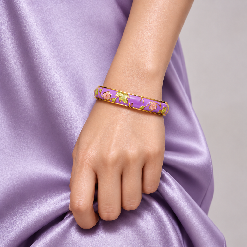 A close-up of a person's hand and wrist wearing a purple enamel bangle bracelet with gold floral detailing, set against a lavender satin fabric background.