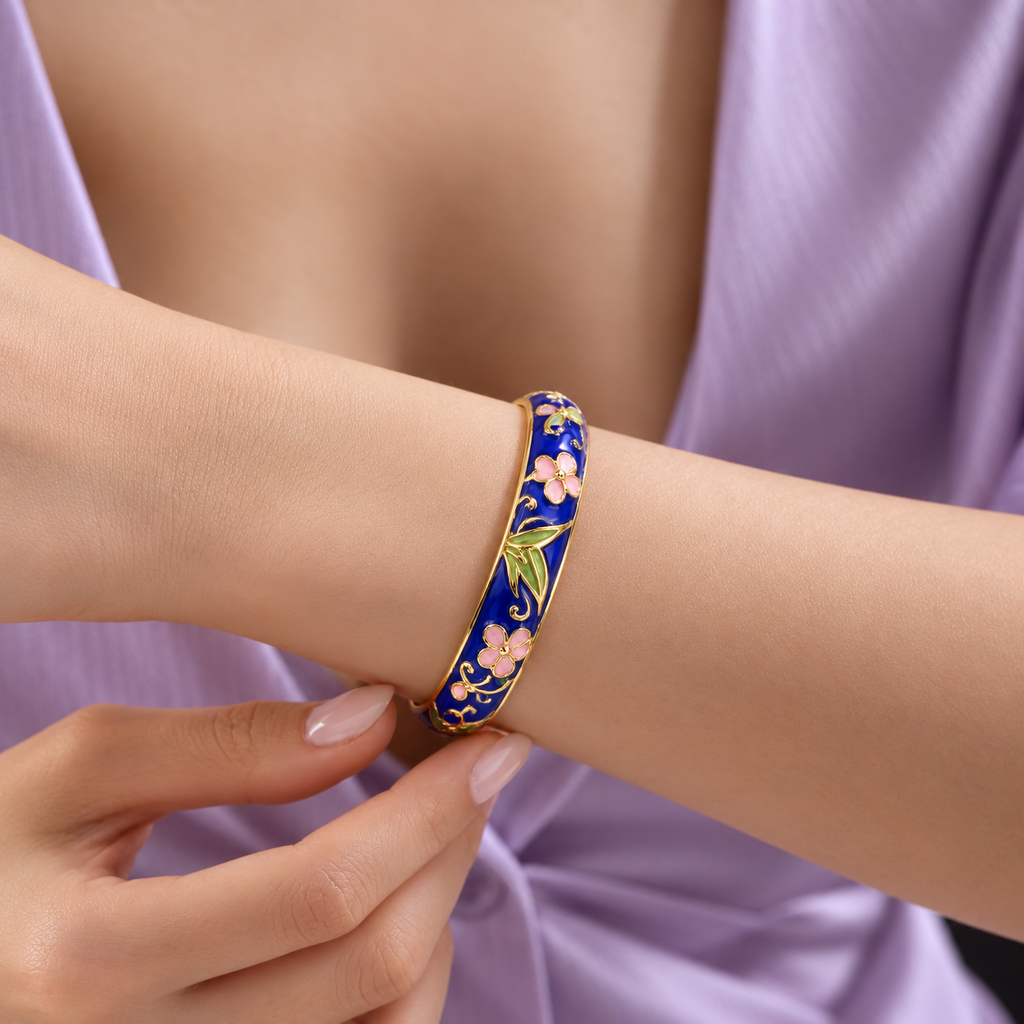 A close-up of a person's wrist wearing a gold-plated bangle bracelet decorated with a traditional Chinese clover pattern featuring a deep blue background, pink flowers, and green leaves.