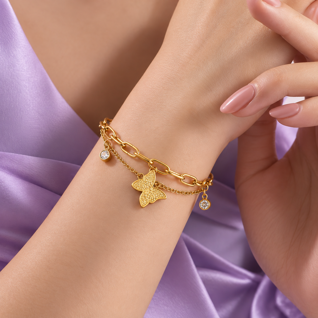 A close-up of a woman's wrist wearing a delicate gold chain bracelet with a butterfly charm and small dangling crystal accents, styled with lavender satin fabric in the background.