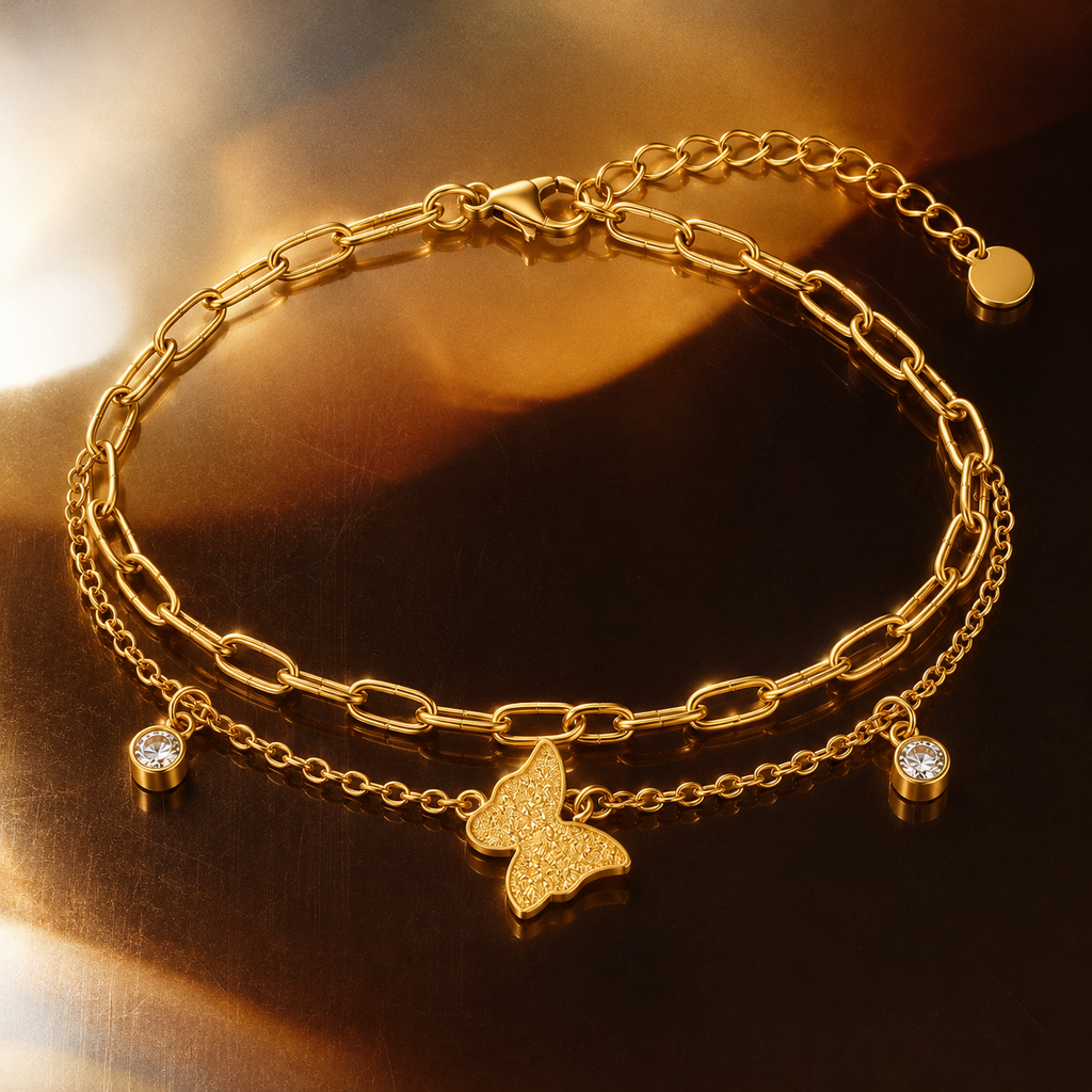 A gold-toned double-layer bracelet featuring a paperclip chain, a delicate chain with a butterfly charm, and two small bezel-set clear stones, displayed on a reflective dark brown surface.