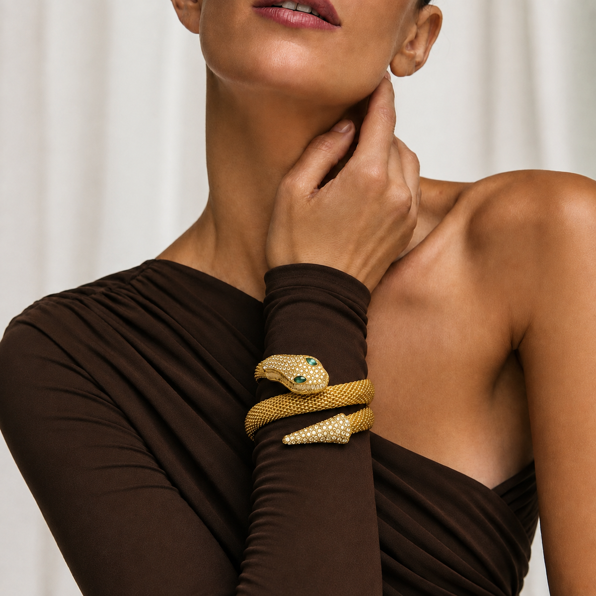 A close-up lifestyle shot of a model wearing a sculptural gold snake cuff bracelet on her wrist, paired with a dark brown draped top against a soft white curtain background.