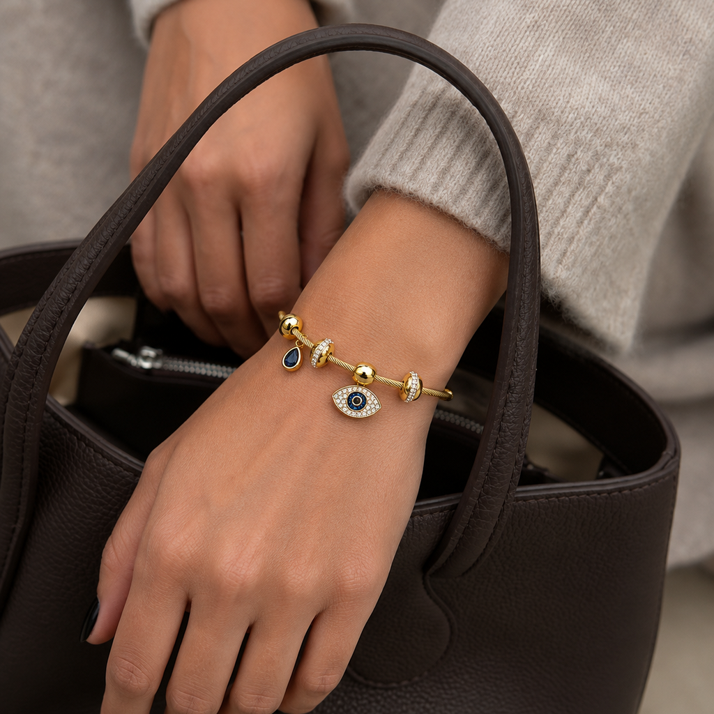 A close-up of a person's hand and wrist wearing a gold charm bracelet with evil eye and blue stone charms, holding a dark brown textured leather handbag.